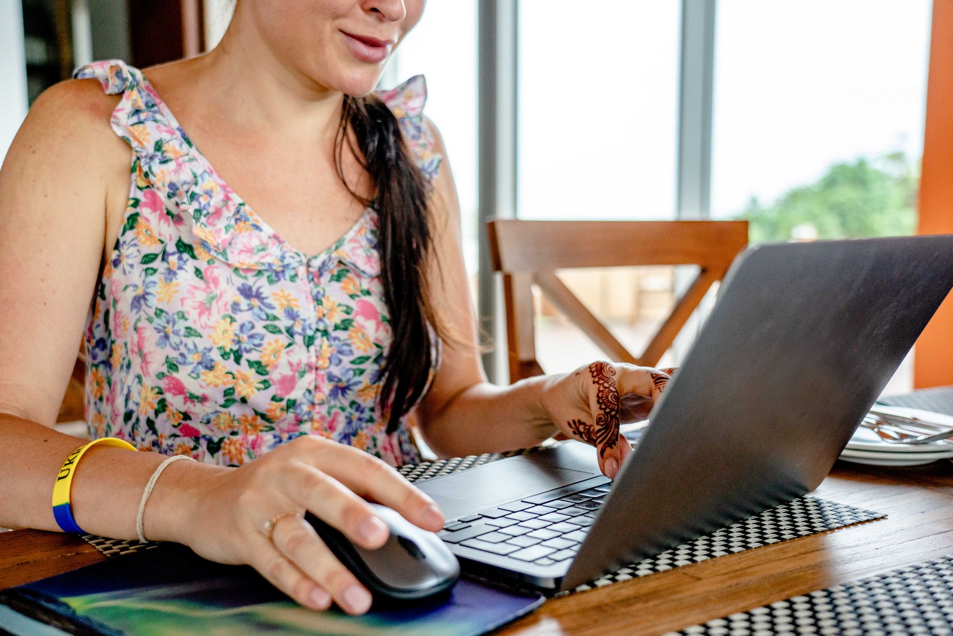 Kobieta pracująca zdalnie na laptopie w kwiecistej sukience, z henną na dłoni.